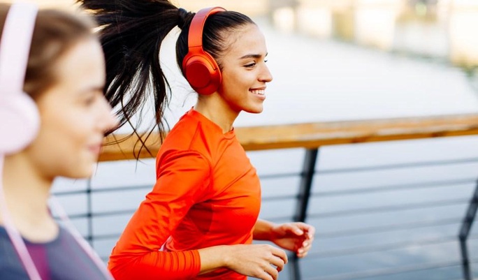  Duas mulheres correndo ao ar livre em um calçadão próximo à água. A mulher em destaque sorri, veste uma camiseta laranja e usa grandes fones de ouvido vermelhos.